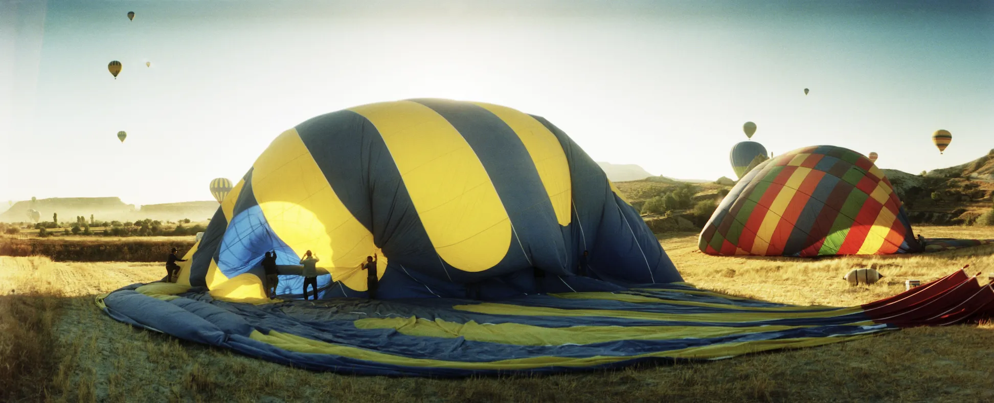 A hot air balloon collapsed and deflated on the ground at dawn, with crew gathering the fabric while other balloons fly overhead