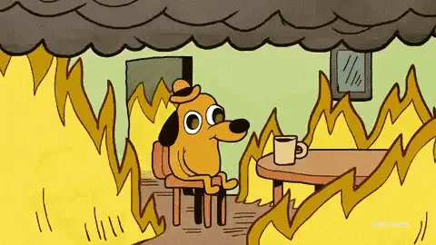 The animated This is fine dog from KC Green's comic: a cartoon dog in a hat sits at a table with a mug of coffee, smiling, while flames spread across the room around it